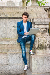 Fototapeta na wymiar European college student studying in New York. Wearing blue blazer, jeans, sneakers, a young guy staying at quiet corner on campus, working on laptop computer, reading, thinking. Working Anywhere..