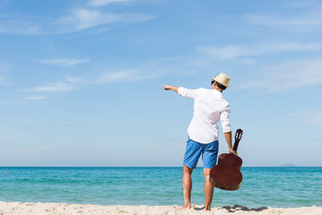 Back view of man holding guitar and standing on the beach with blue sea and blue sky bacjground. Travel relax vacation concept..