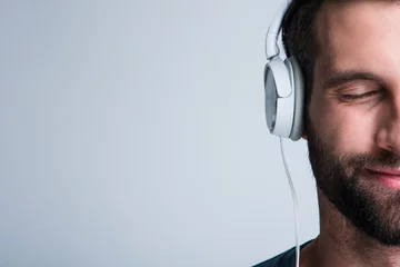 Poster Perfect song. Part of portrait of handsome young man listening to music in headphones with smile while standing against white background © MARIIA