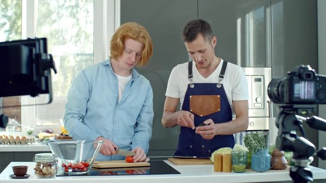 Medium shot of professional chef in apron cutting avocado and chatting with young man with ginger hair slicing tomatoes while filming cooking show in modern kitchen