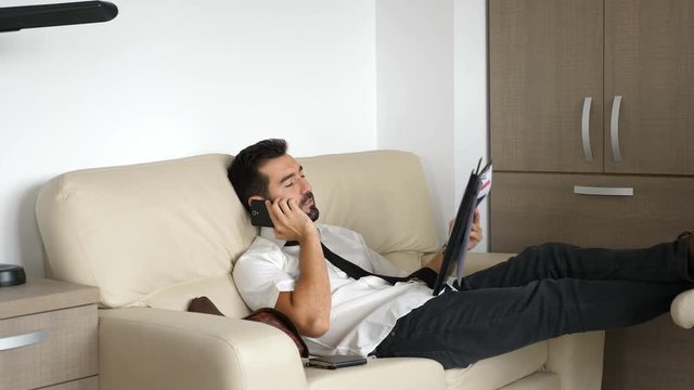 Revealing shot of tired businessman on the couch talking on the phone and examing some documents and charts