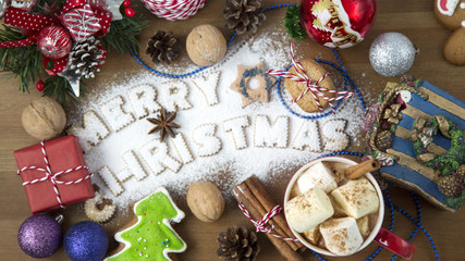 Hot Christmas Beverage Cocoa and Marshmallow in Small Cup with Lettering Text Merry Christmas from Cookies Gift Boxes Cord Sparkles Fir Branch Nuts Cinnamon Sticks Anise on Wooden Background. Top View