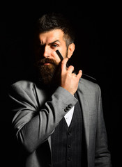 Man with long beard holds blade for razor near cheek.