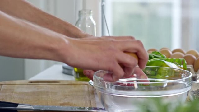 Tilt down of young male professional cook cracking eggs into clear bowl standing on kitchen table with fresh vegetables lying on plate