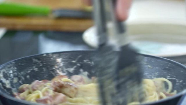 Close up of unrecognizable cook with spatula tongs stirring spaghetti frying in hot pan with cheese sauce and bacon, then serving it on plate