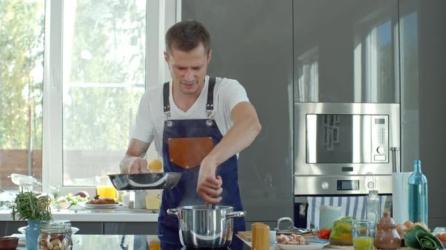 Medium shot of male chef in apron standing at kitchen countertop and taking spaghetti out of boiling water with tongs, then frying them in pan and talking to camera
