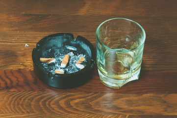 Glass with alcohol drink and dirty black ceramic ashtray full of cigarette butts on the wooden table surface close up selective focus