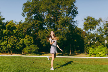 Young pretty girl with long brown hair dressed in light clothes staying on green lawn grass in the park on trees background. Summer sunny time.