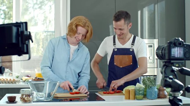 Medium shot of professional chef in apron and young man with ginger hair slicing tomatoes and chatting during filming of cooking show in modern kitchen