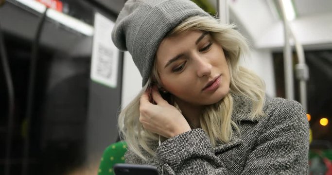 Portrait of cute girl wearing on headphones and listening to music on mobile phone in public transport. City lights background