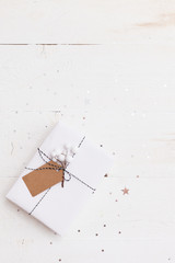 Top view on elegant Christmas gift wrapped in white gift paper, Christmas tree decorations on white wooden background with sparkling stars. New Year, holidays and celebration concept