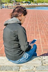 Young Woman Using her Smartphone while Sitting on a Bench along a Waterfront Path. Baltimore, MD.