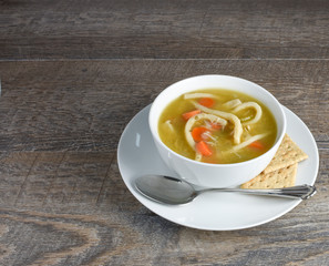 Overhead view of homemade chicken noodle soup with spoon and whole grain saltine crackers on rustic weathered wood background.  Ingredients noodles, carrots, celery, chicken, broth.