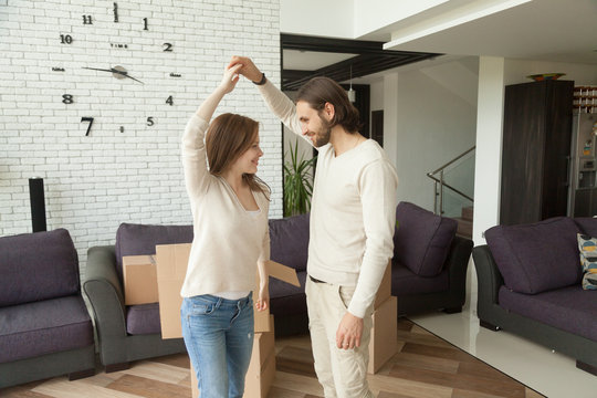 Happy couple dancing in new home after moving in, smiling homeowners having fun packing boxes in living room interior, man and woman feel excited about own apartment, young family enjoying relocation