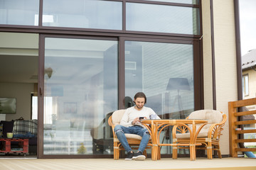 Young man sitting on chair at terrace of own modern house with glass wall, relaxing with cup of tea using mobile phone, attractive smiling guy enjoying spending time outdoors holding cellphone alone