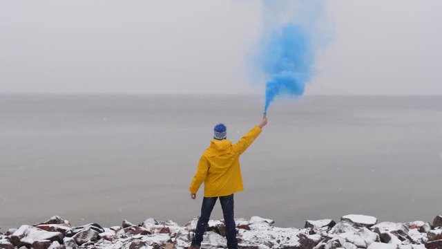Rear view of man with blue smoke smoke in yellow coat in front of frozen sea. Snow falling in foreground.