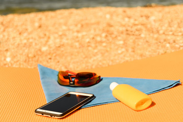 red Sunglasses lying next to a smartphone on a microfiber towel next to the sunscreen on the travel rug on the beach
