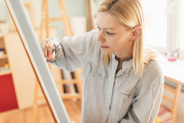 Fototapeta premium Beautiful blonde woman artist drawing with a brush and pain in her art studio