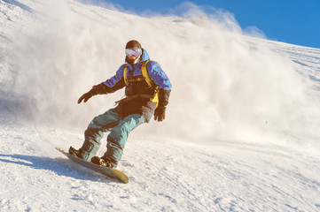 Freeride snowboarder rolls on a snow-covered slope leaving behind a snow powder against the blue sky