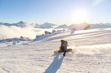 A snowboarder in a ski mask and a backpack is riding on a snow-covered slope leaving behind a snow...
