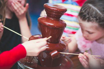 Vibrant Picture of Chocolate Fountain Fontain on a children kids birthday party with a kids playing around and dipping marshmallows and fruits into the fountain
