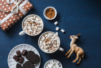 Obraz na płótnie Canvas Christmas inspiration table - hot chocolate with marshmallows, cookies, gift box, christmas ornament reindeer on a blue background, top view. Flat lay
