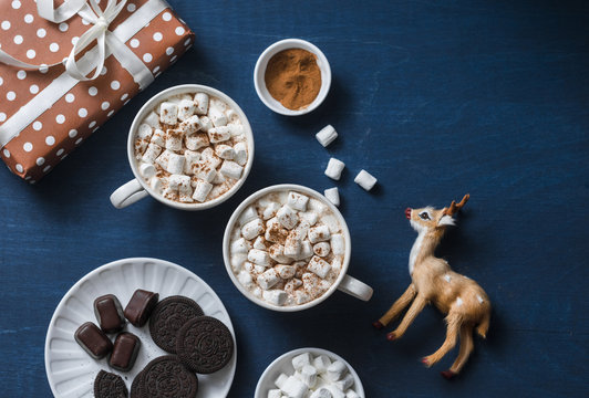 Christmas inspiration table - hot chocolate with marshmallows, cookies, gift box, christmas ornament reindeer on a blue background, top view. Flat lay