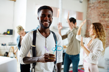 Happy African-american guy with drink looking at camer aon background of dancing friends