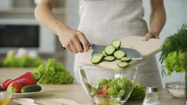Cooker cutting fresh cucumber into slices and adding it into bowl with salad