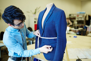 Young tailor measuring waist of blue jacket on mannequin in his workshop