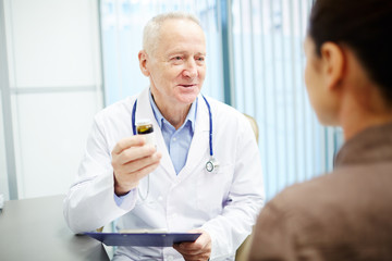 Mature doctor in whitecoat describing new pills in pill-bottle to his patient