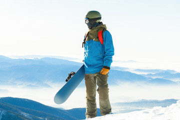 Young snowboarder standing at the very top of a mountain and holding his snowboard with one hand