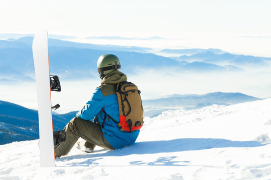 Young snowboarder sitting next to snowboard thrusted into snow and looking at a  beautiful scenery before him