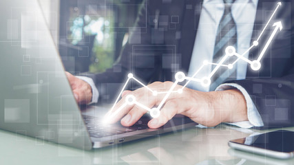 Obraz premium Hand of Businessman typing on laptop with smartphone and graph connection shape rise up in home office or co working space background .Concept of workplace using mobile technology.