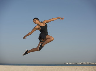 Fototapeta na wymiar Beautiful Inter-racial Black Asian Fitness Model does an explosive dance moves wearing headphones on a white sandy beach on a clear blue sunny morning. 