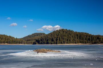 Winter landscape. Frozen lake, pine forest. Bulgaria, Rhodopes mountains, Shiroka Polyana lake. Rocky island in front.