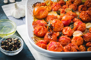 Roasted Italian cherry tomatoes with garlic, herbs and olive oil on dark blue stone background. Selective focus, close up.