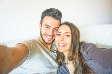 Happy young couple taking a selfie with mobile smart phone camera in the living room embracing on sofa at home - Friends making self portrait on the couch - People, relationship, lifestyle concept