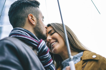 Happy couple kissing under an umbrella in a rainy day - Handsome man kiss his forehead girlfriend...