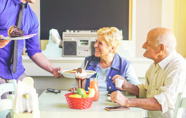 Happy couple of seniors enjoying together a sweet lunch in a bar restaurant - Waiter serving muffin and pancakes to a table of couple of pensioner - Elderly, lifestyle and food concept