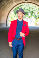 Fototapeta premium Businessman traveling in New York. Dressing in red blazer, blue collarless shirt, pants, wearing Fedora hat, a young guy walking through arch gate, confidently looking forward. .
