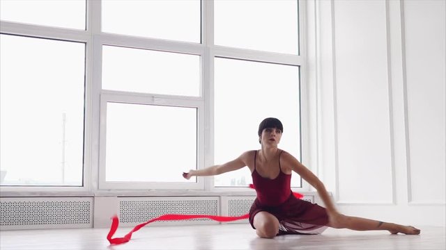 Beautiful young woman is spending her time practicing moves with a ribbon. Sitting on the floor doing rhythmic gymnastic.