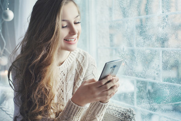 Fototapeta premium Indoor close up portrait of young beautiful happy smiling girl using her smart phone. Model wearing stylish clothes. Day light from window. Female beauty, fashion concept. Copy space for text. 