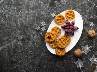Homemade soft waffles for breakfast with frozen berries of organic northern cranberries on a white oval ceramic dish on a gray grunge background with silver Christmas decor and a garland. Top View.