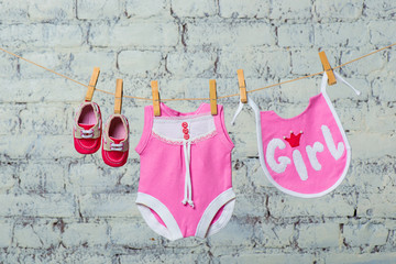 Children's pink body, bib and red shoes, dry on a rope against a white brick wall.