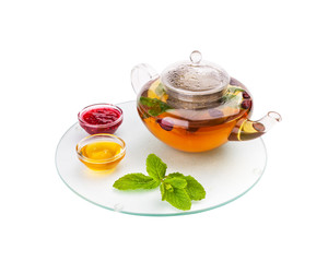 The glass teapot full of hot herbal tea with a dogrose costs on a glass opaque tray with saucers, with honey and jam, and mint leaves.
