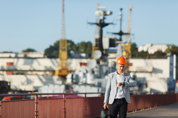 Handsome young unshaven successful business man in gray suit and protective construction orange helmet holding case, looking time on watch, walking in sea port against cargo ship and crane background