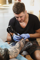 Obraz premium Professional tattooer artist doing picture on hand of man by machine black ink from a jar. Tattoo art on body. Equipment for making tattoo art. Master makes tattooed in light studio.