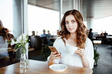 the nice business lady speaks by smartphone in street cafe. the girl is dressed in a white blouse and holds a cup of coffee in hand.