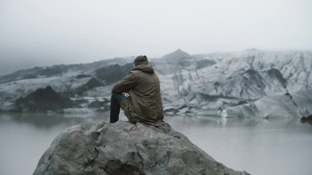 Back view of young man sitting on the volcanic rock alone and looking on the glaciers in Ice lagoon in Iceland.